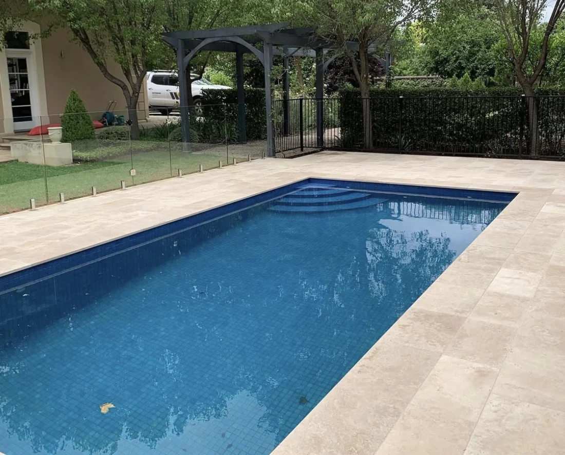 Completed blue swimming pool with pale travertine paving and garden surrounds