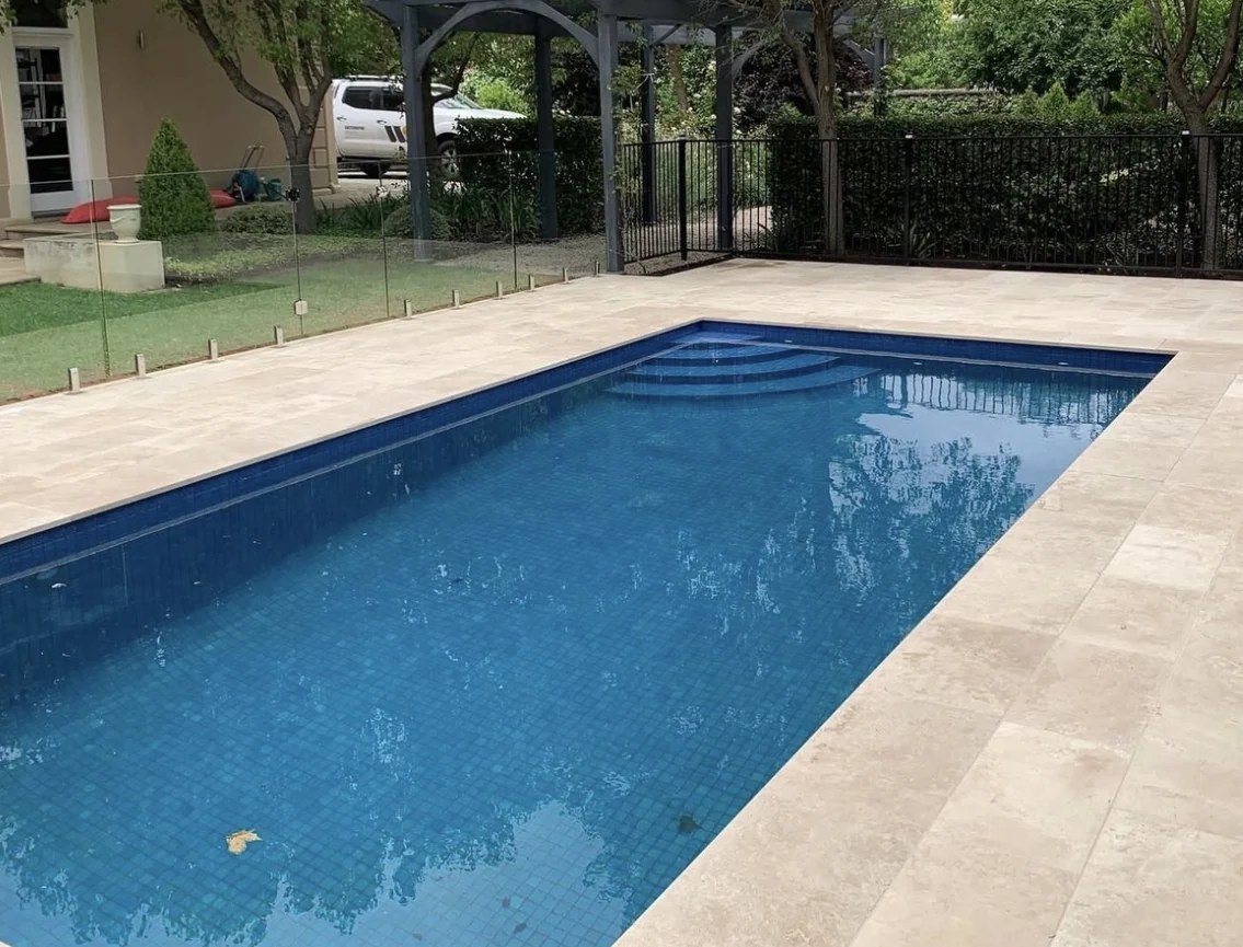 Completed blue pool framed by pale stone paving, glass fencing and established trees